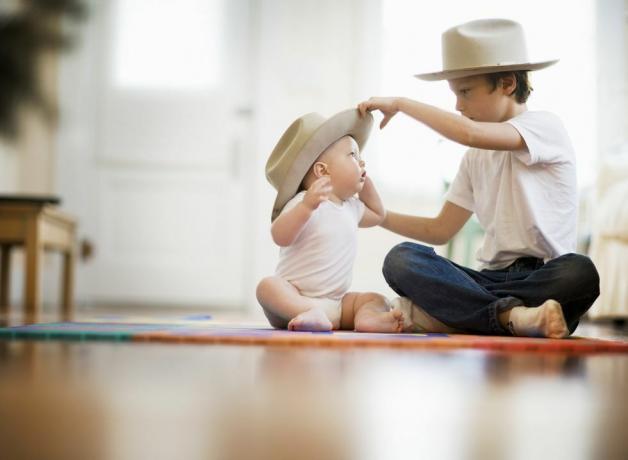 दो भाइयों का चयनात्मक फोकस दृश्य, एक नौ साल का, एक आठ महीने का, जोड़ा फर्श पर क्रॉस लेग करके बैठता है, बड़ा लड़का बच्चे के सिर पर मैचिंग टोपी लगाता है