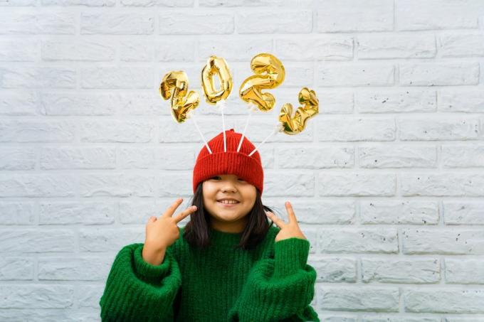 हरे रंग के स्वेटर में खुश एशियाई बच्ची, टोपी में 2023 नंबर वाले सुनहरे गुब्बारों के साथ, सफेद ईंट की दीवार की पृष्ठभूमि, कैमरे की ओर देख रही लड़की, मुस्कुरा रही है