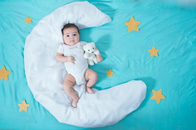 सुंदर लैटिन बच्ची, दो महीने की, चंद्रमा के आकार की एक सफेद चादर पर लेटी हुई है जिसके दोनों ओर पीले तारे हैं और उसके नीचे एक नीली चादर है।