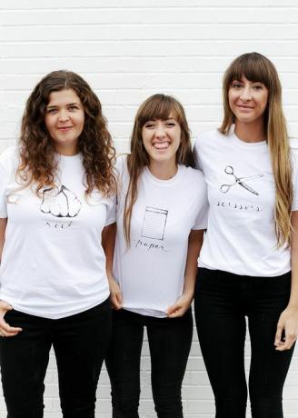 सफेद टी शर्ट में तीन महिलाएं, या तो रॉक, कागज या कैंची के साथ शर्ट पर लिखा और चित्रित किया गया है