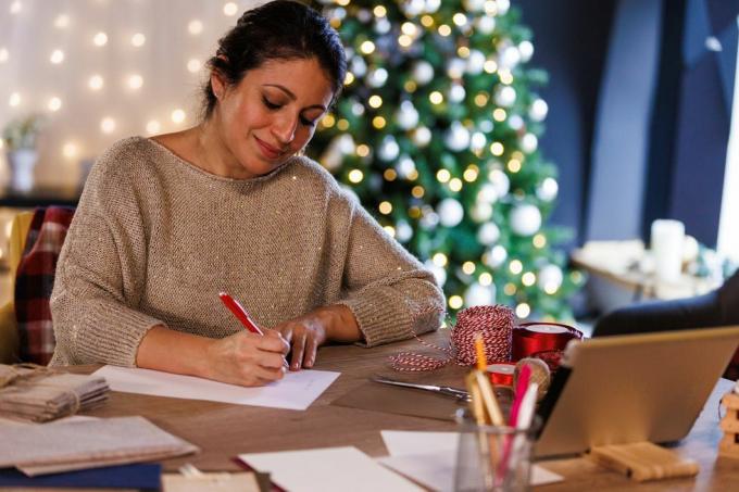 चमचमाते क्रिसमस ट्री के सामने मेज पर बैठी एक खुश मध्य वयस्क महिला का चित्र, जो प्रियजनों के लिए क्रिसमस कार्ड और पत्र लिखते हुए मुस्कुरा रही है।