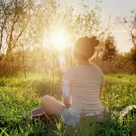 योग मुद्रा में बैठी युवा महिला प्रकृति में सूर्यास्त का आनंद ले रही है