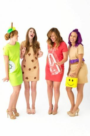 "लंच लेडीज" के रूप में कपड़े पहने छोटी पोशाक में चार हंसती हुई महिलाएं, एक पीले स्माइली फेस लंच बॉक्स के साथ